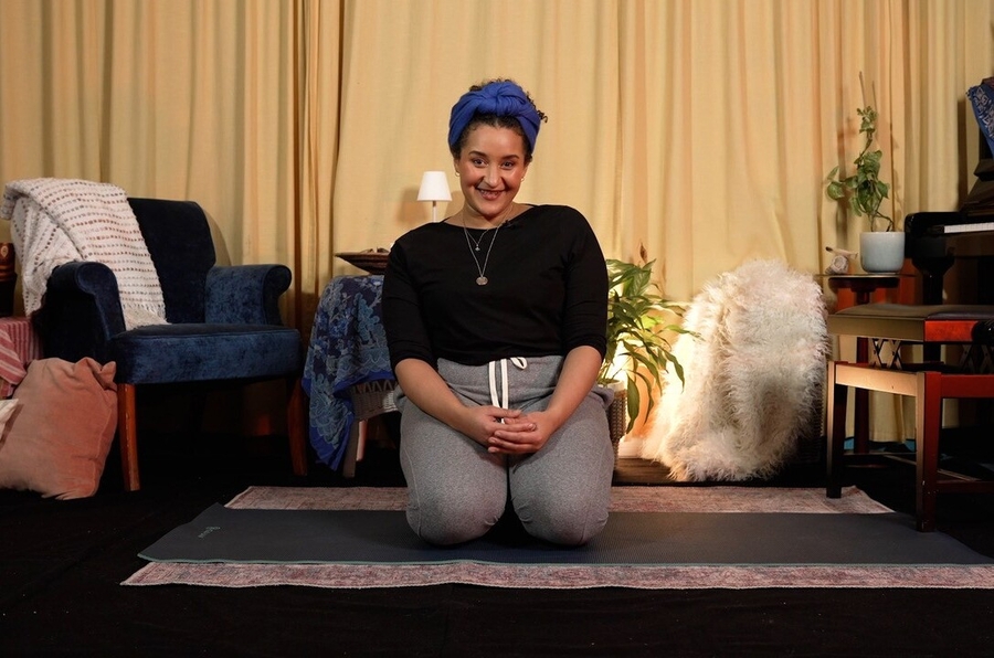 A portrait of Lea Shaw. She is smiling to the camera and kneeling on a yoga matt.